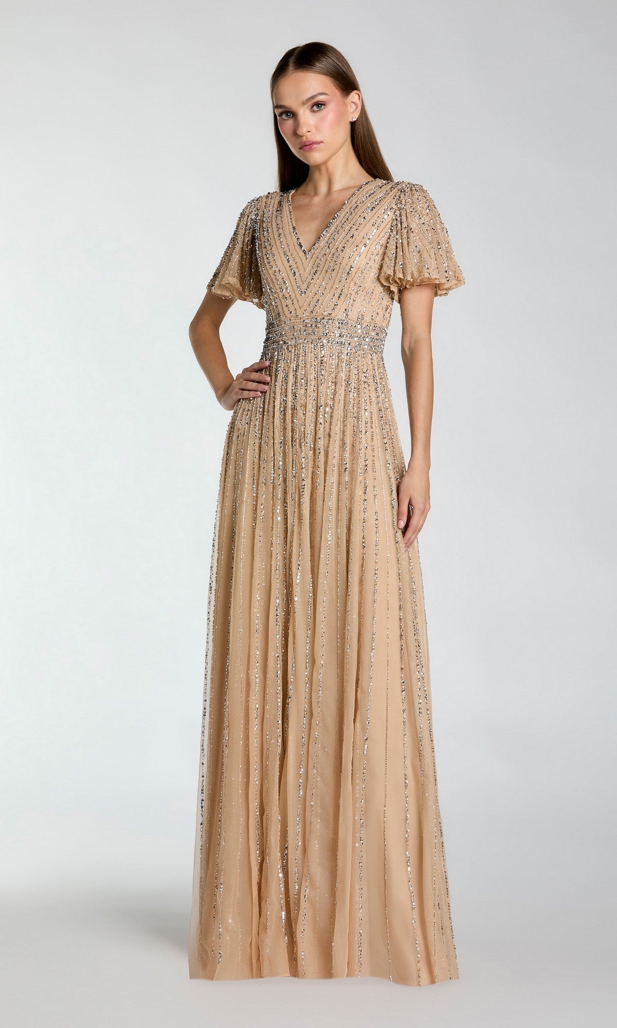 A woman wearing the Mac Duggal 5713 long formal dress in nude silver, featuring short sleeves and allover silver embellishments, stands against a plain light background.